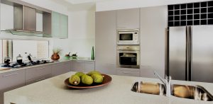 Counter top made in ceramic seen closely with fruits on a brown color clay dish next to the modern  silver faucet attached to the sink, the silver refrigerator and the wall oven with pantry cupboards next to the gas stove